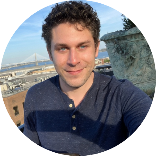 Man with curly brown hair and blue eyes smiling on a rooftop, with a bridge in the background.