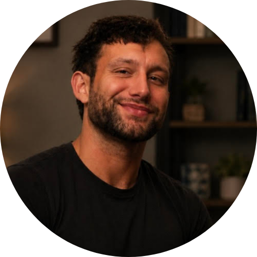 Smiling man with dark hair and beard wearing a black shirt, indoors in a cozy setting.